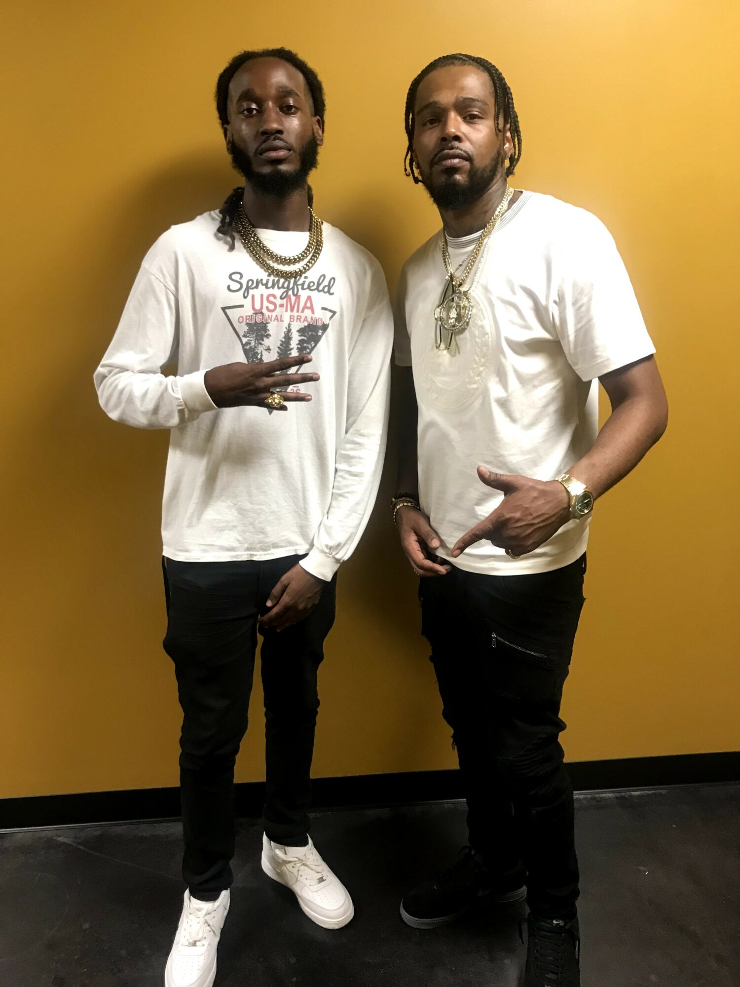 Two men with long hair and beards stand against a yellow wall, wearing casual white shirts and black pants.