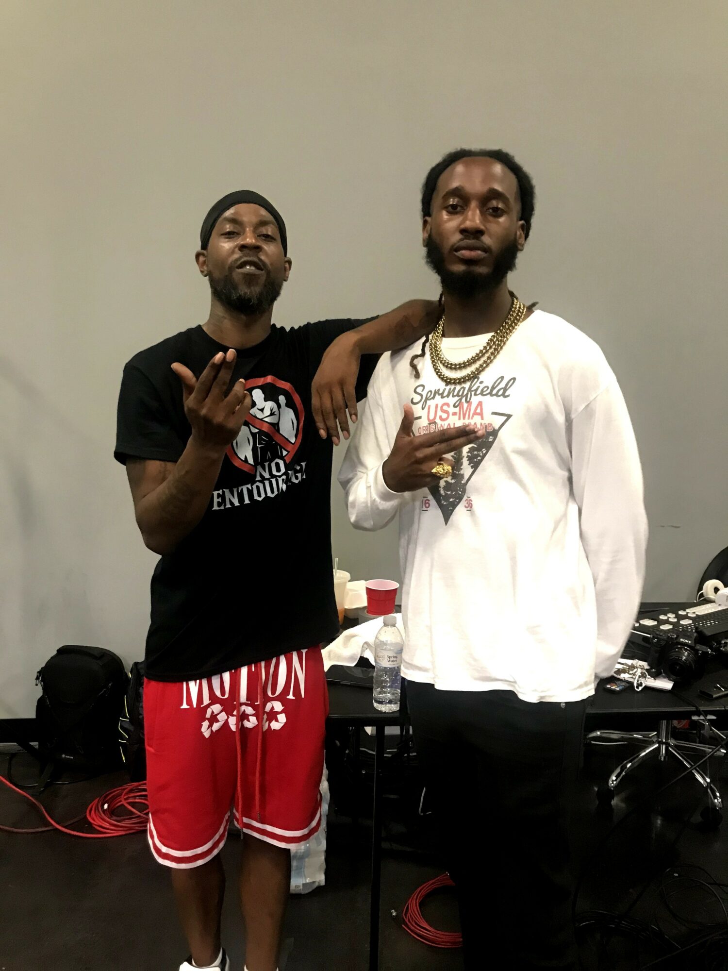 Two men standing side by side, one with arm around the other, in a room with a table and equipment behind them.