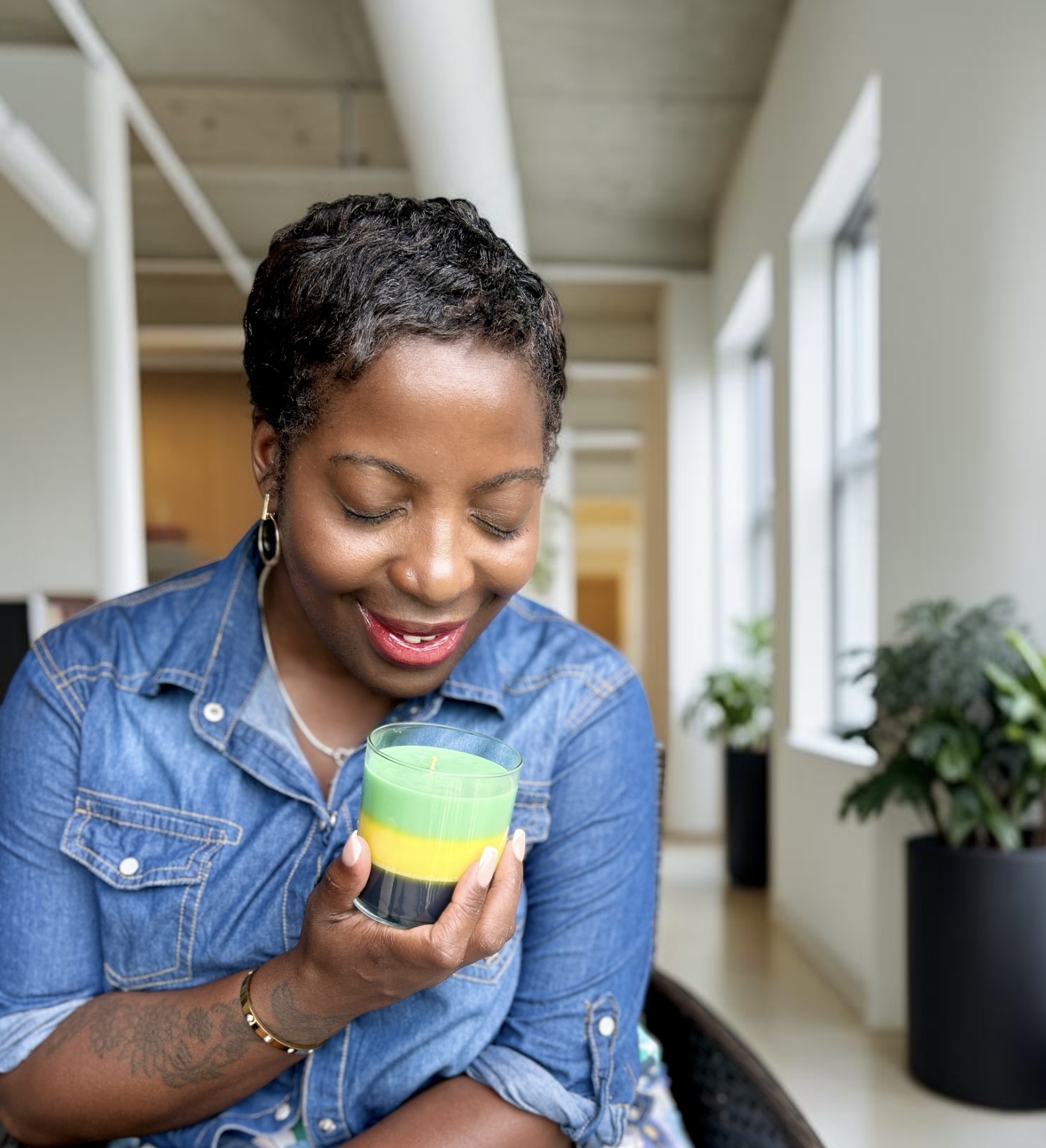 Woman with short curly hair smiling and holding a small container, indoors with large windows and potted plants.