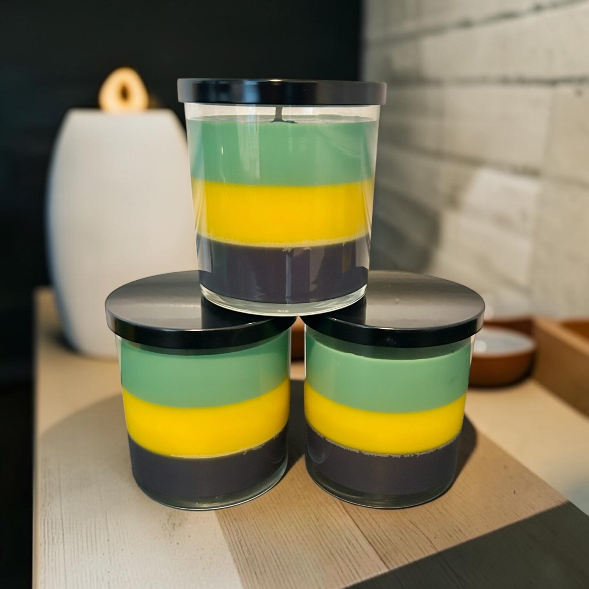 Three layered candles with green, yellow, and purple layers, stacked on a wooden surface, with a white vase in the background.
