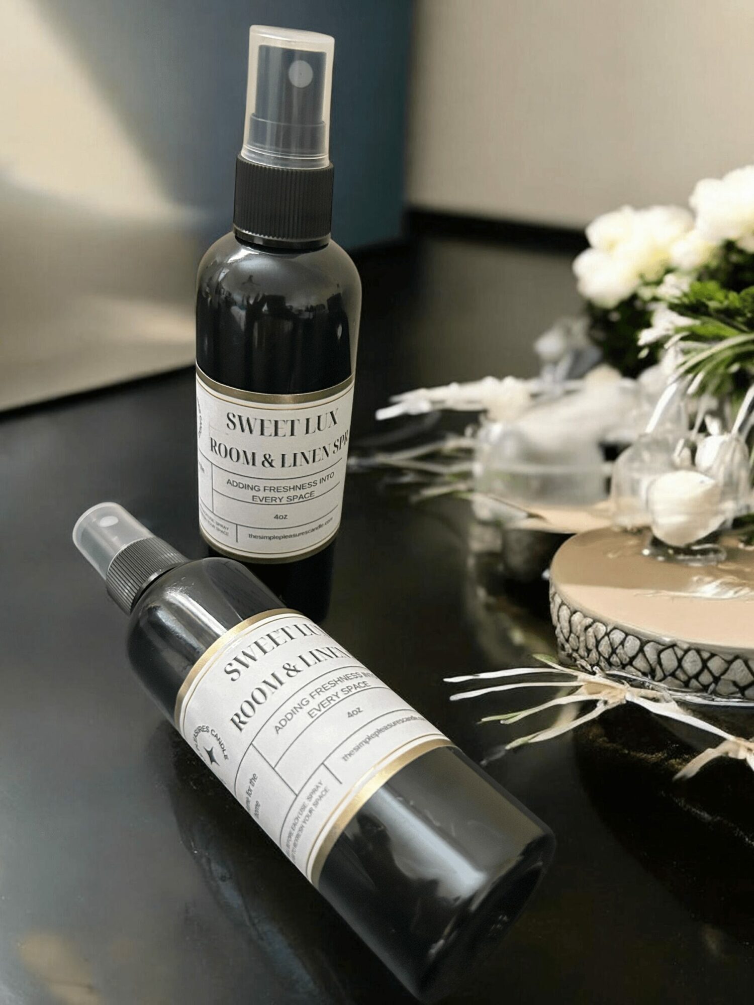 Two black spray bottles with white labels on a black surface, with flowers and decorative items nearby.