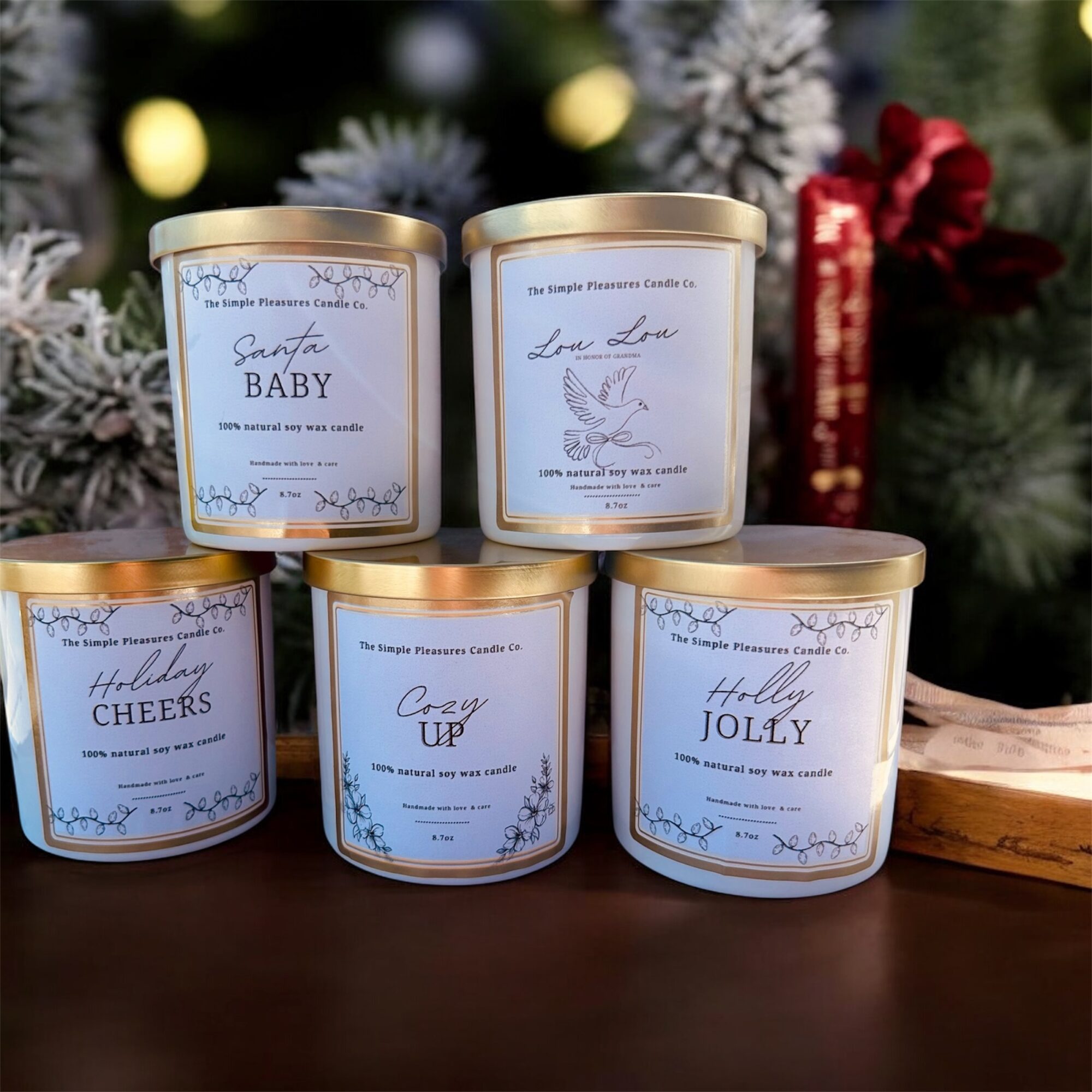 Five candles with gold lids, decorated with holiday-themed labels, placed on a dark surface with Christmas decorations in the background.