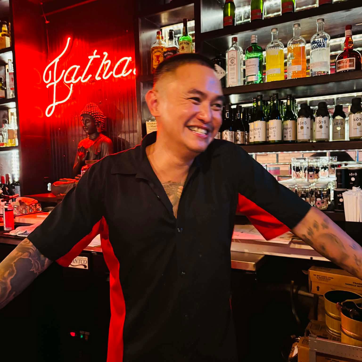 Smiling person with tattoos behind bar with bottles and a red neon sign in the background.