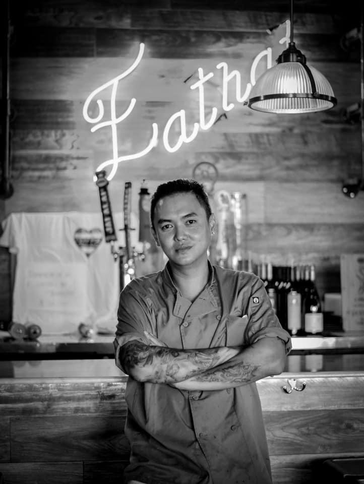 Person with tattoos and short hair standing in front of a bar with bottles, a neon sign, and hanging lamp, in black and white.