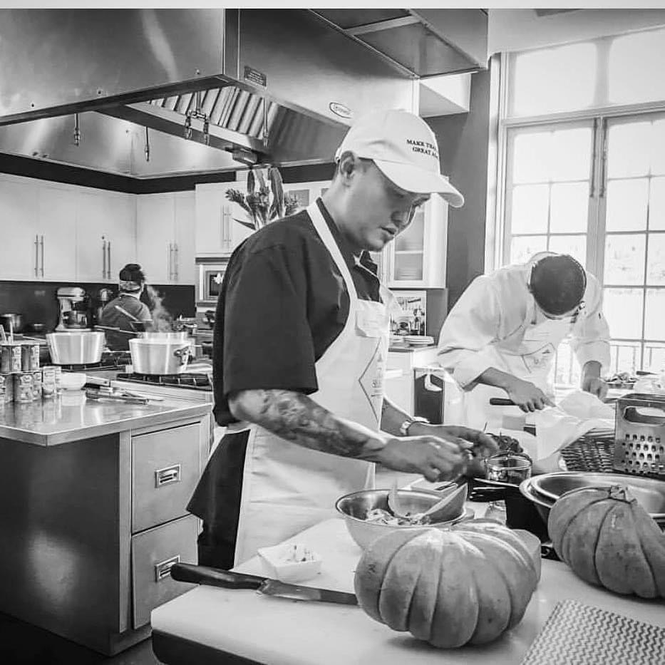 Two people working in a kitchen, one wearing a cap and apron, preparing food with pumpkins on the counter.