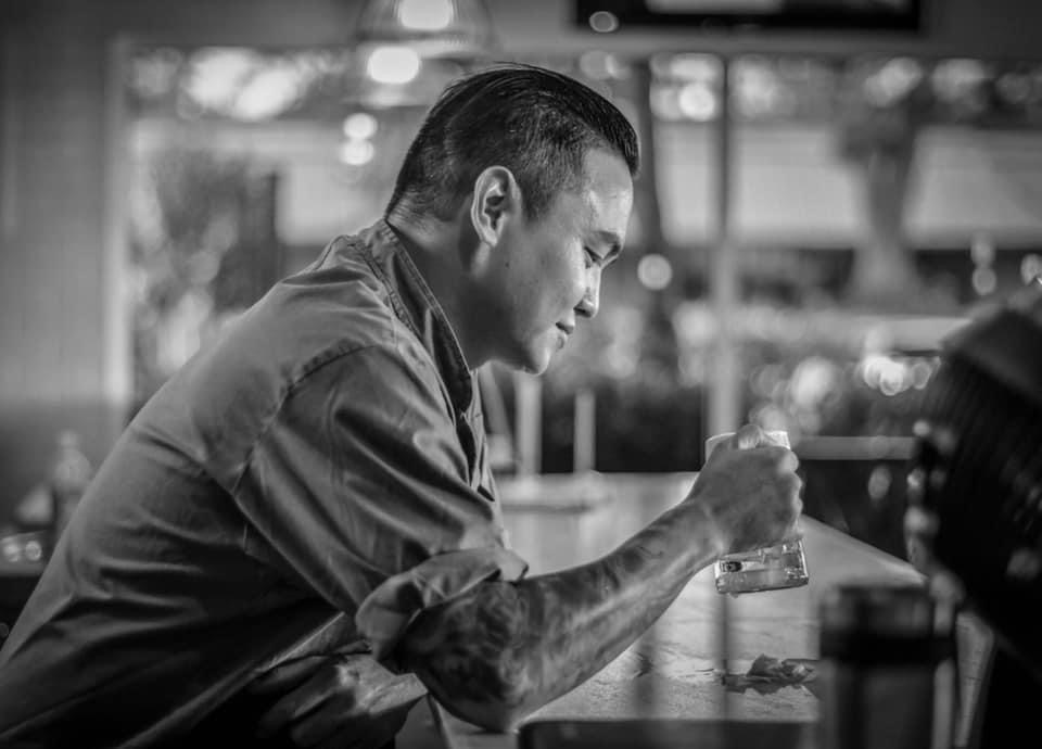 Man sitting at a table holding a glass, with a blurred background, in black and white.