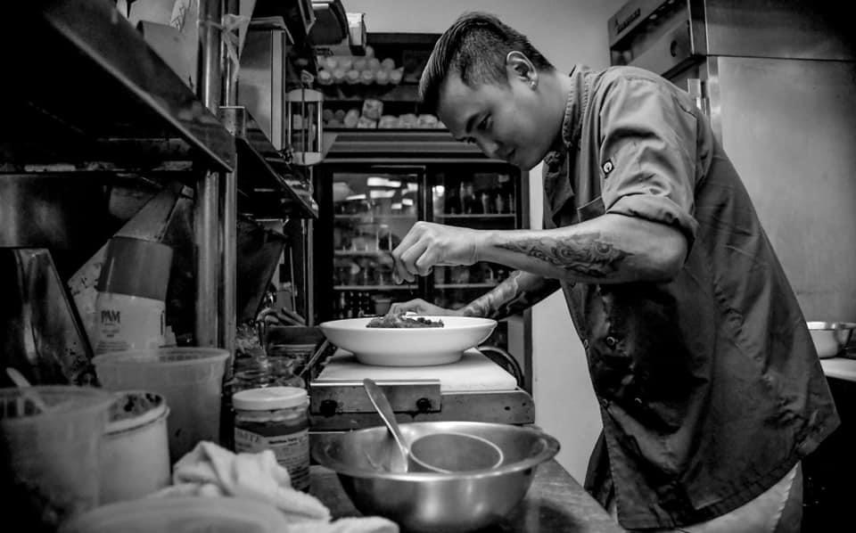 Person preparing food in a kitchen, focused on a bowl, surrounded by kitchen utensils and ingredients.
