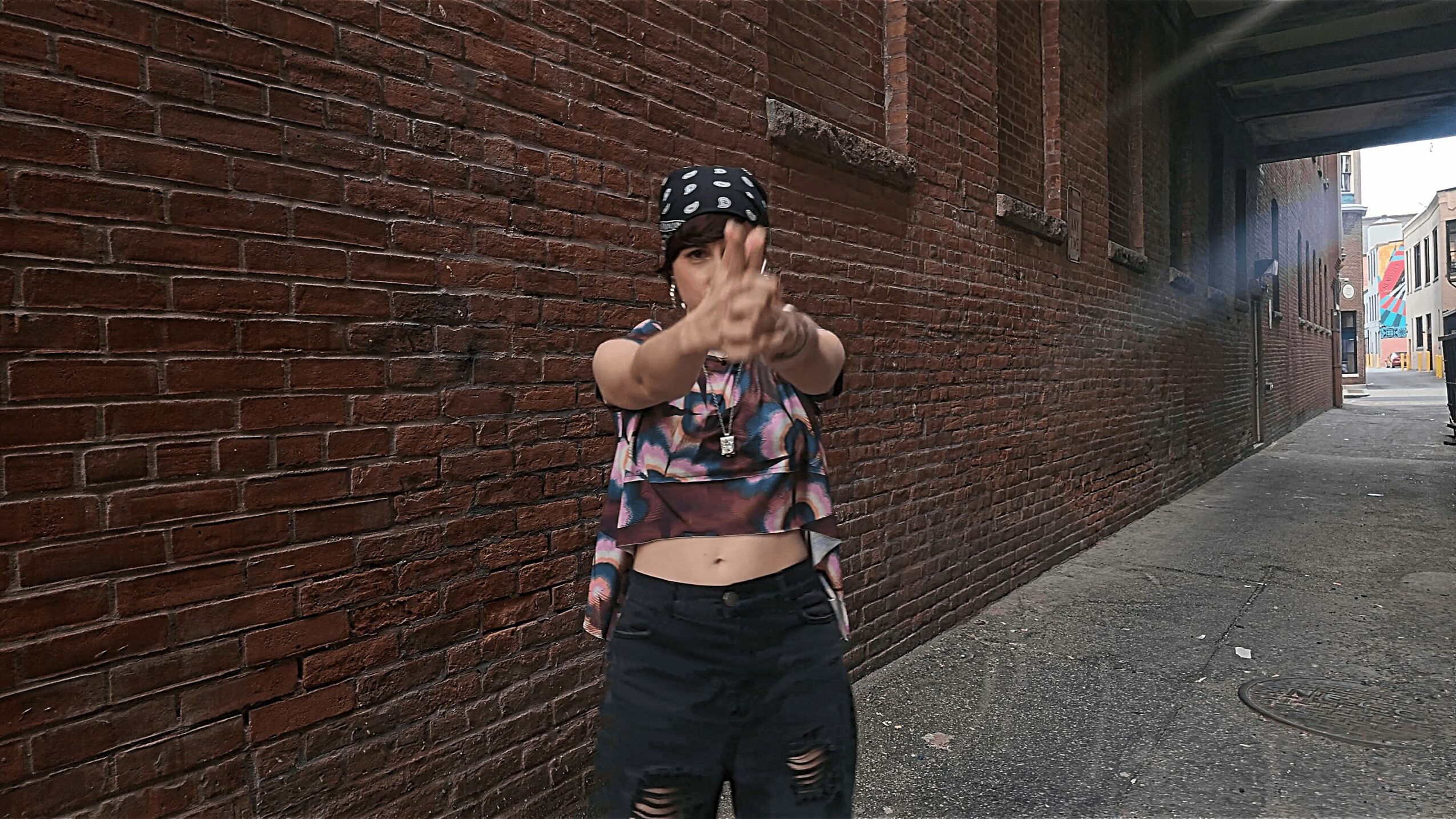 Person standing against a brick wall, wearing a bandana, crop top, and ripped jeans, pointing towards the camera.