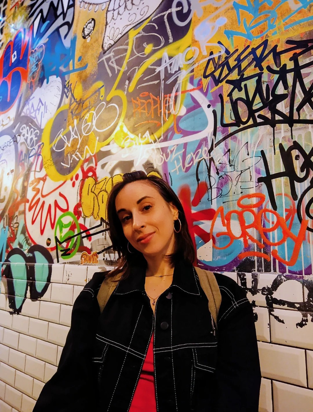 Young woman with dark hair and earrings stands in front of a colorful graffiti wall.