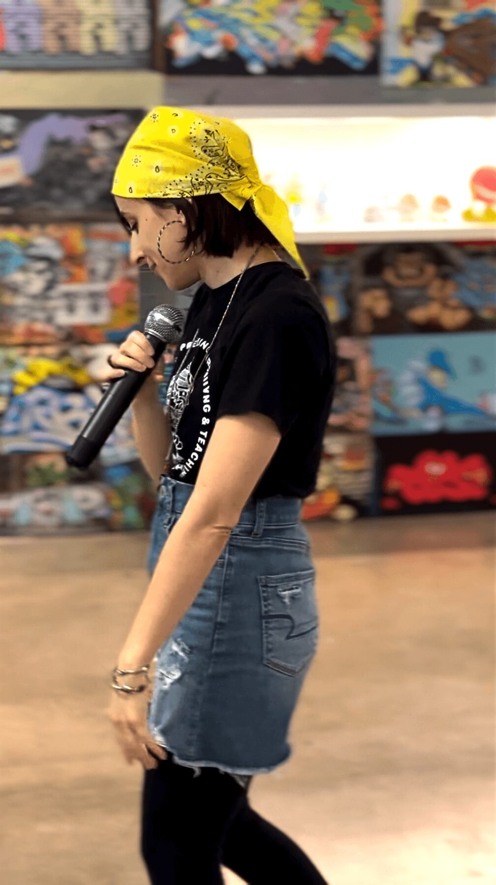 Person wearing a yellow bandana, black T-shirt, and denim skirt holding a microphone in a store.