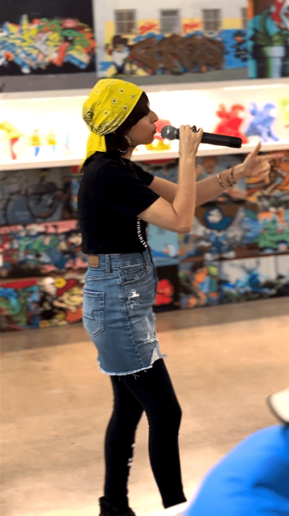Girl wearing yellow hat, black shirt, denim shorts, and black tights, holding a microphone in a colorful store.