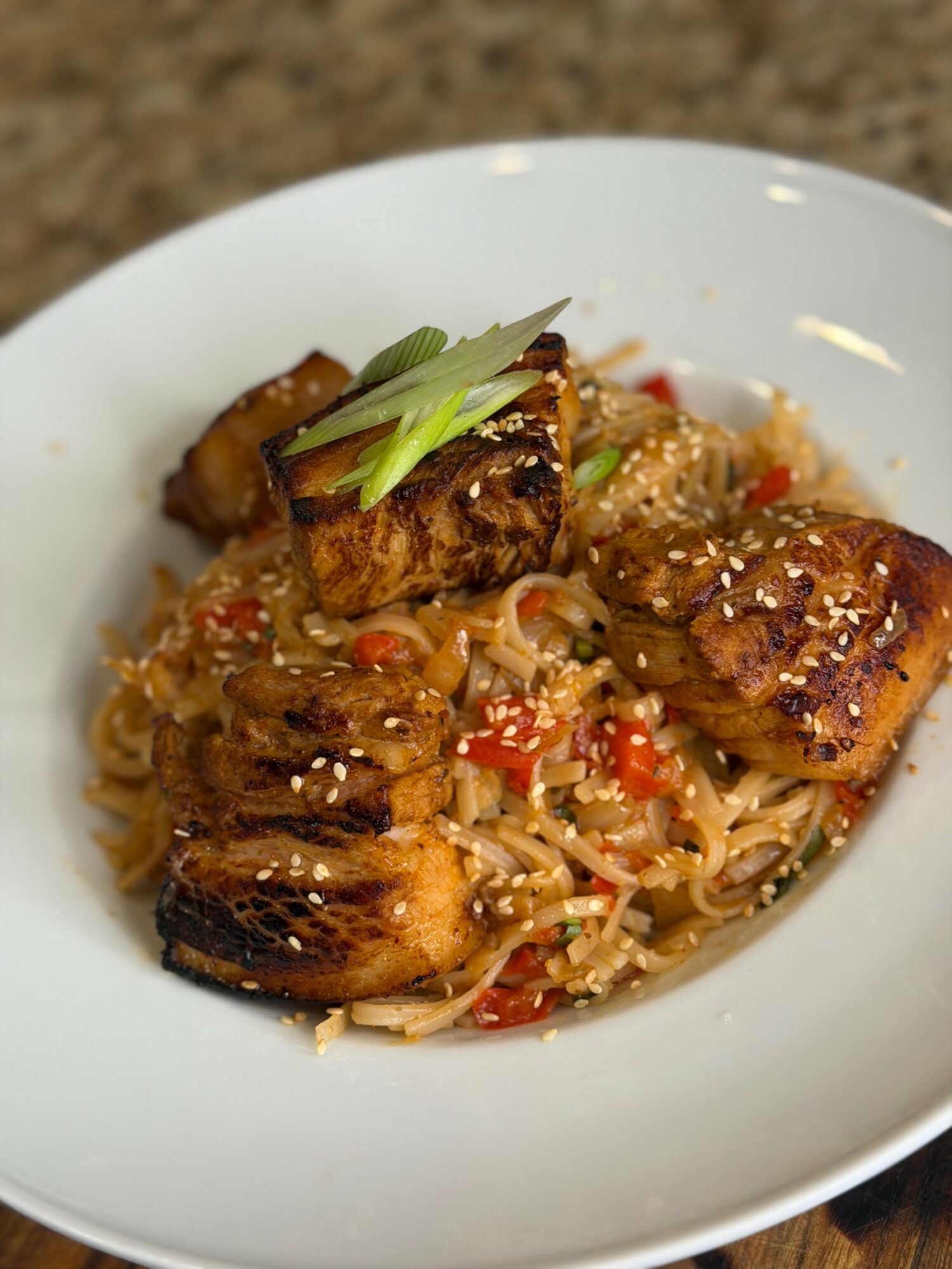 Noodle dish with grilled fish pieces, garnished with green onions, served in a white bowl on a speckled surface.