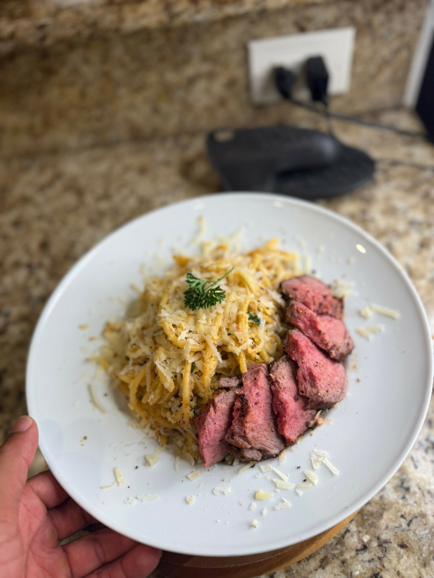 Plate with sliced cooked steak and pasta with cheese on a kitchen countertop.