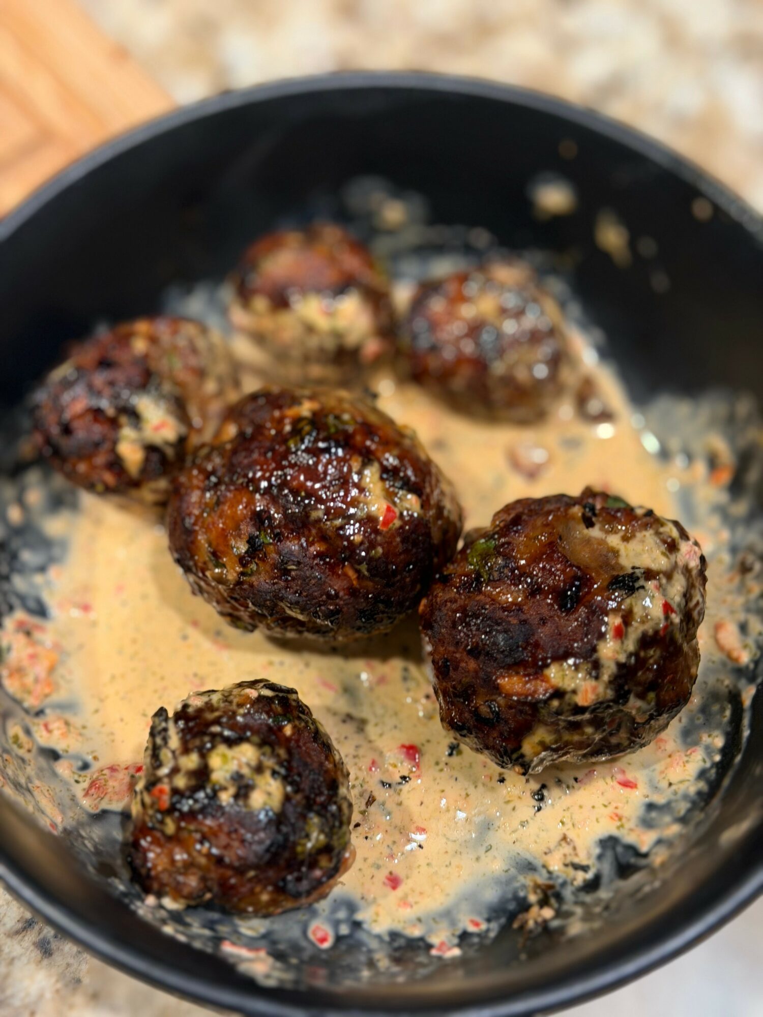 Five cooked meatballs in a black skillet with creamy sauce, some browned and others with bits of cheese or herbs visible.