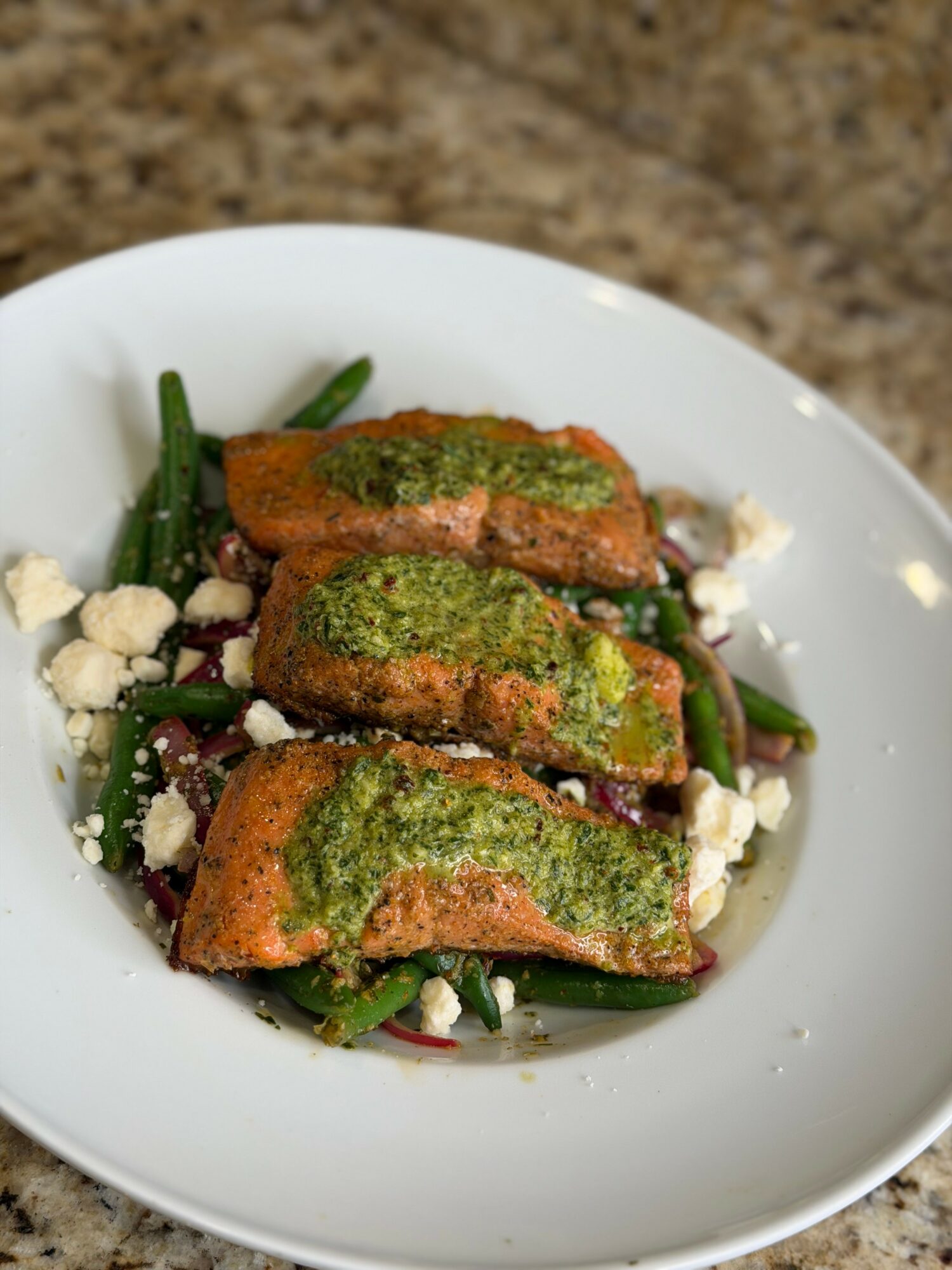 Three pieces of fried food topped with green sauce on a bed of green beans and crumbled cheese in a white bowl.
