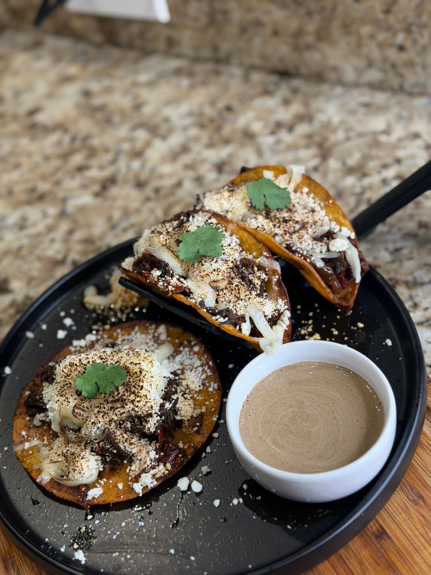 Three caramelized desserts topped with powdered sugar and mint leaves, served with a small cup of sauce on a black plate.