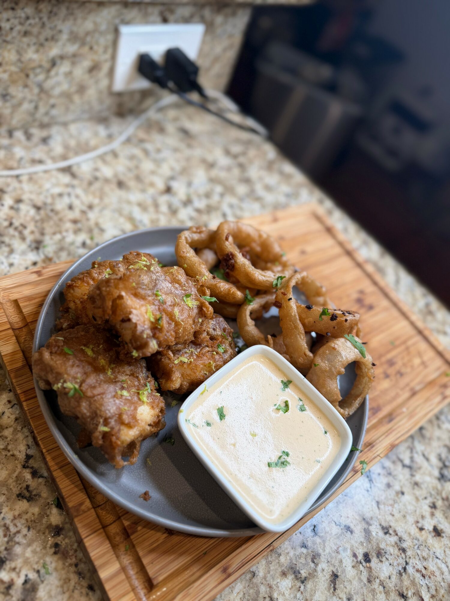 Plate with fried chicken, onion rings, and dipping sauce on a wooden board, on a kitchen countertop.