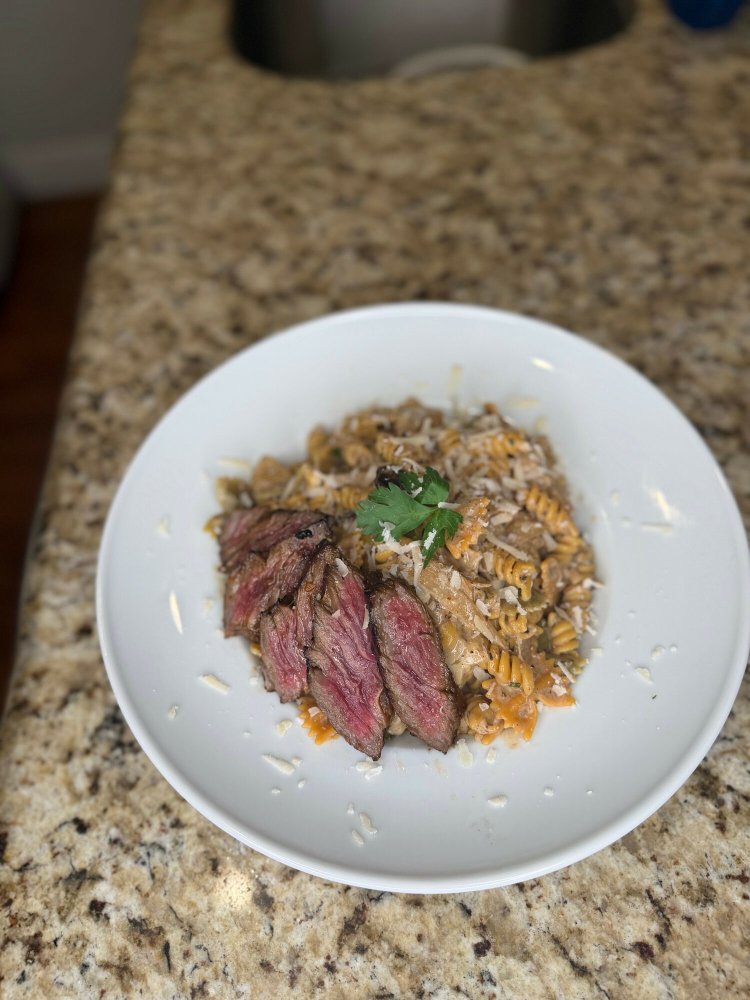 Slices of cooked steak with a side of rice and vegetables on a white plate, garnished with a small green herb, on a granite countertop.