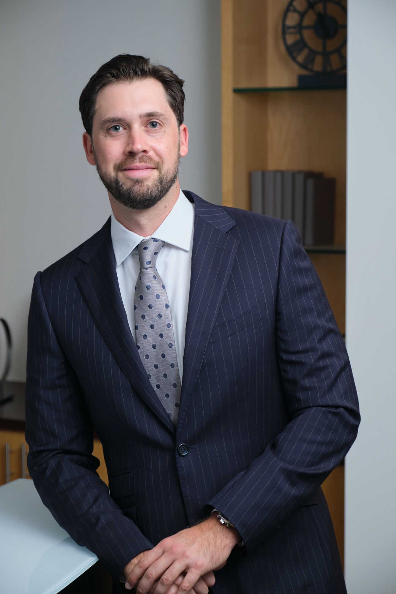 Man in a dark pinstripe suit with a light shirt and patterned tie, standing indoors with a bookshelf in the background.