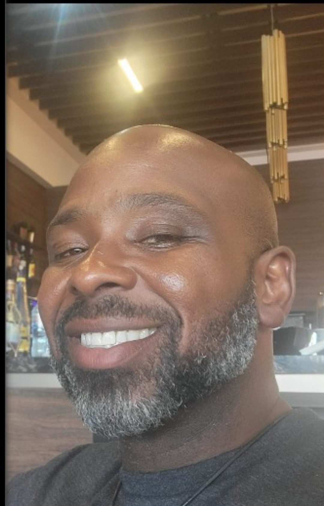 Close-up of smiling man with a beard, indoors with warm lighting and wooden decor in background.