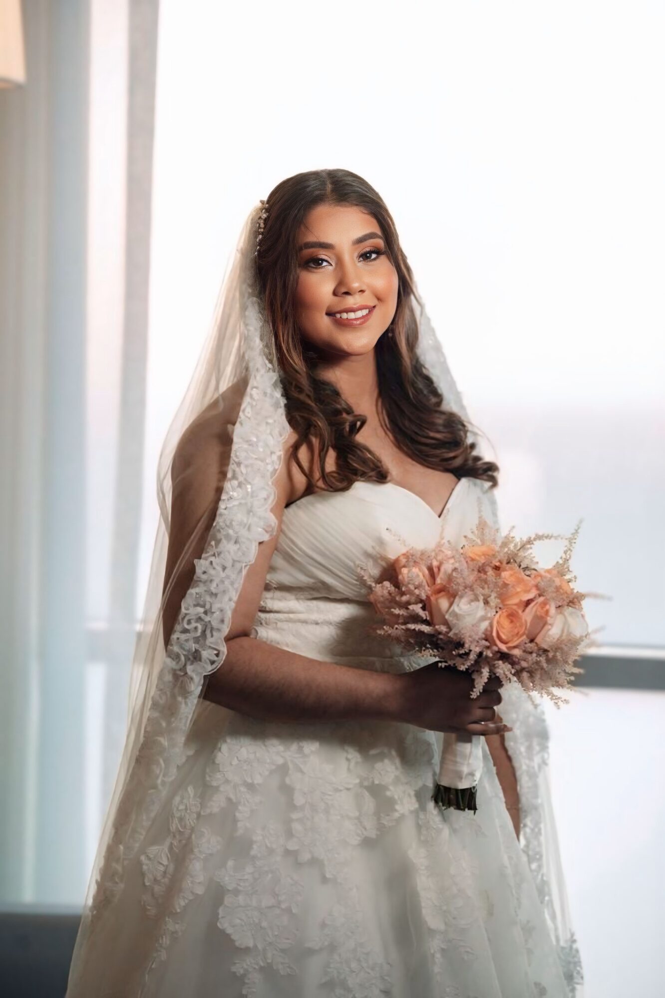 Smiling bride in wedding dress holding a bouquet, standing near a window with natural light, veil draped over her shoulder.