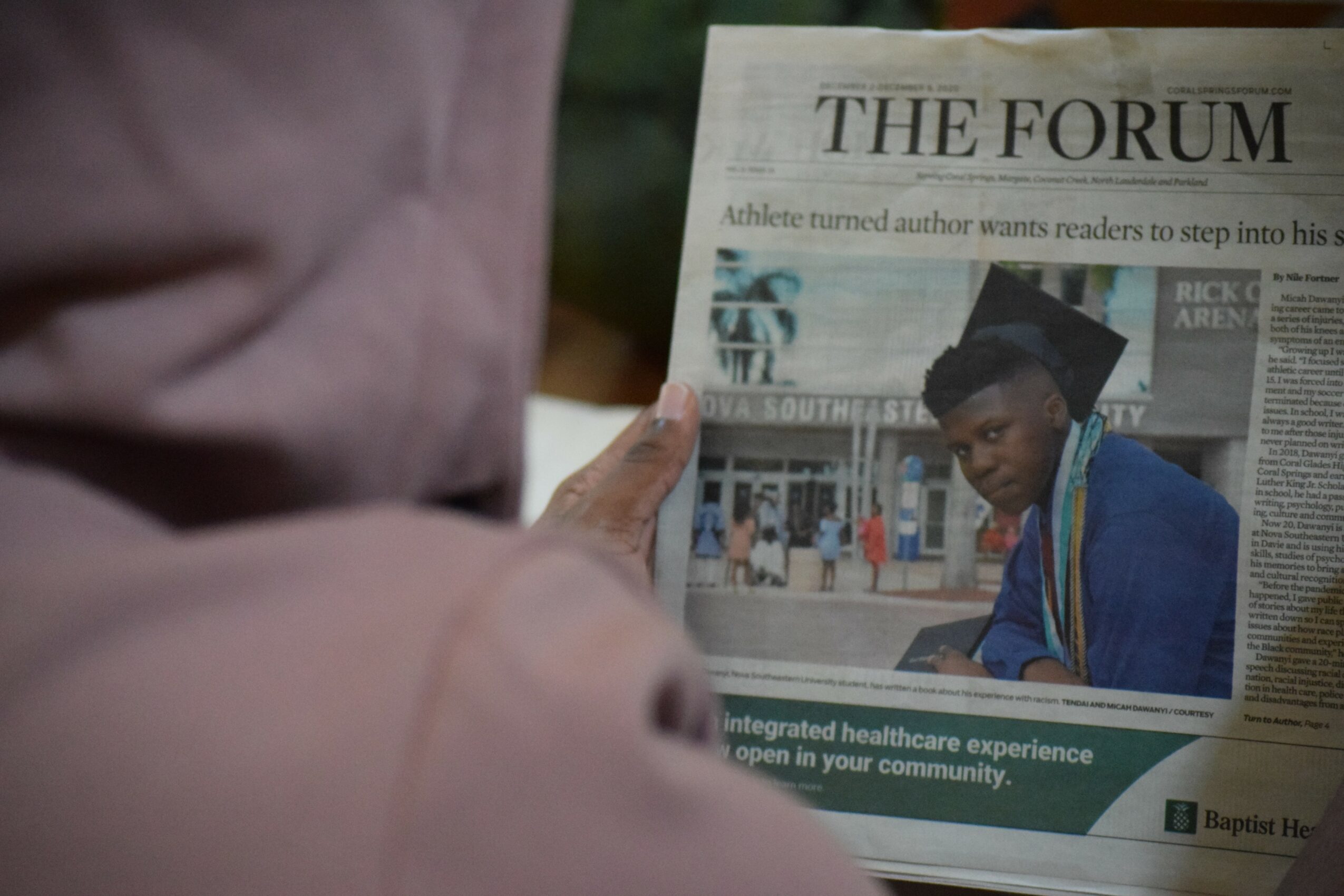 Person reading a newspaper titled 'The Forum' outdoors, with a young man on the front page, holding the paper with both hands.