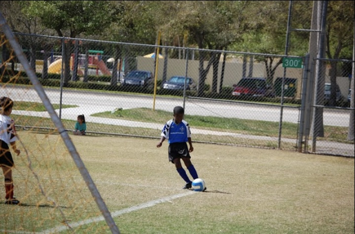 Child in blue and black soccer uniform kicking a ball on a grassy field, with a chain-link fence and parked cars in background.