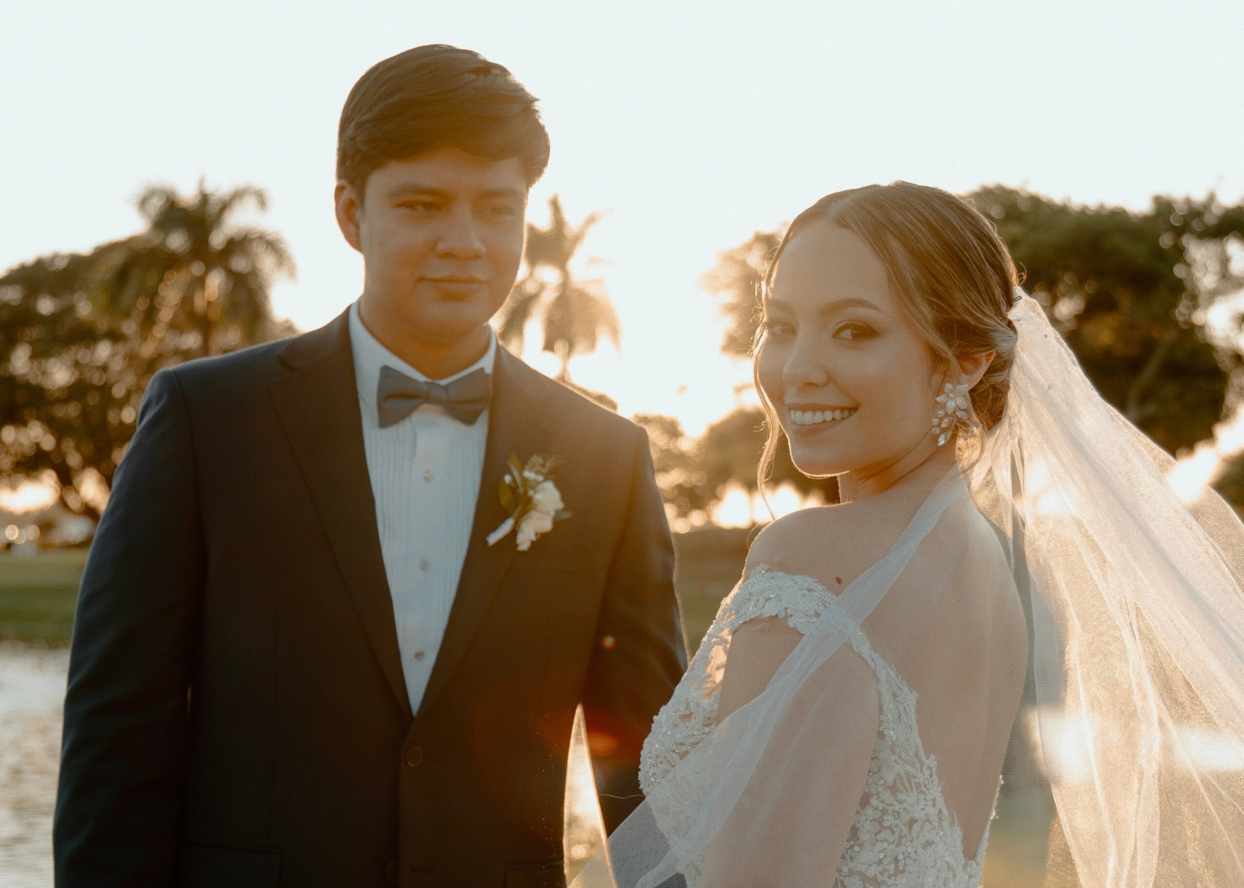 A bride and groom outdoors at sunset, smiling, with trees in the background, dressed in wedding attire.