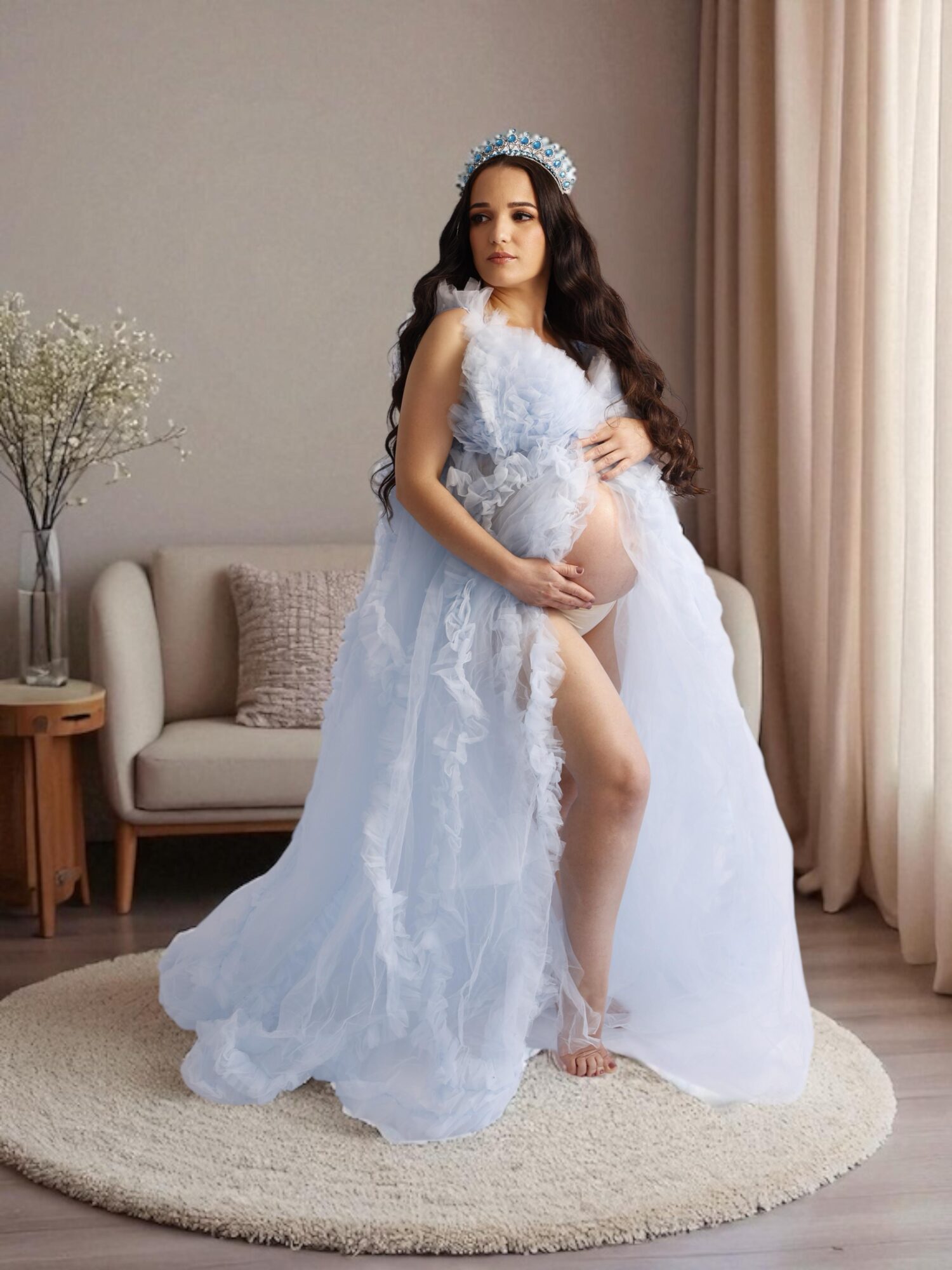 Pregnant woman in a flowing white dress with lace details, standing on a round rug in a living room, holding her belly.