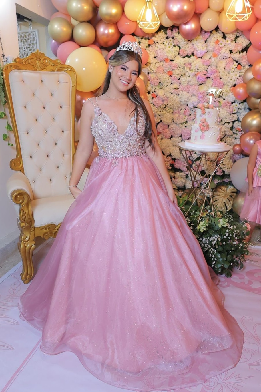 Young woman in pink gown with silver top, standing in front of balloon and floral backdrop, smiling.