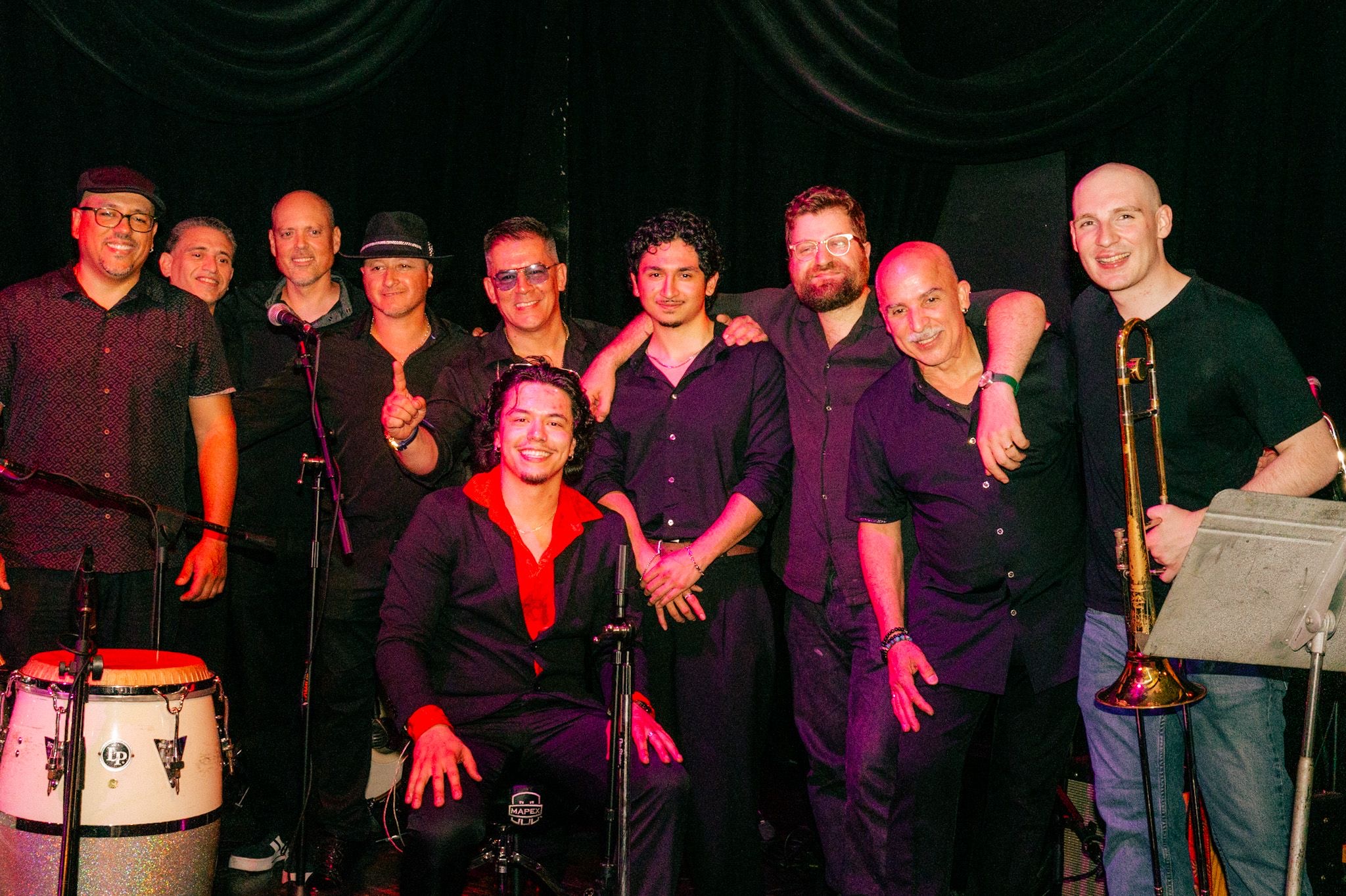 Group of nine musicians on stage, some holding instruments, with a dark background and stage lighting.