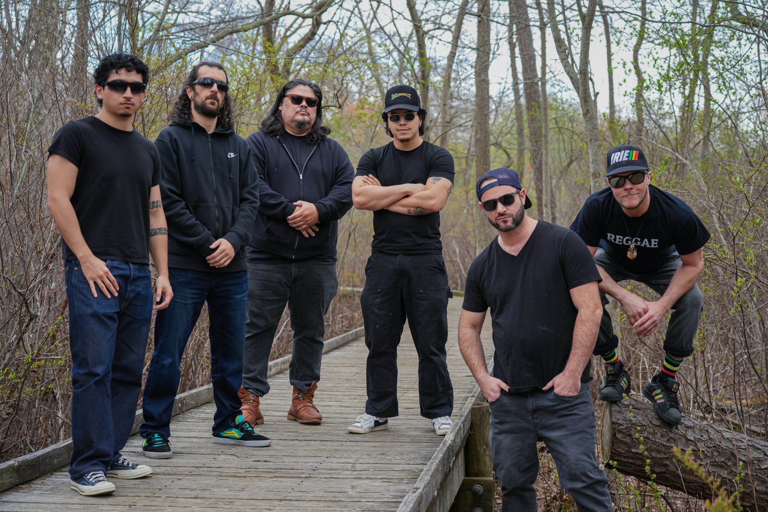 Group of six men standing on a wooden bridge in a wooded area, some wearing sunglasses and casual clothing.