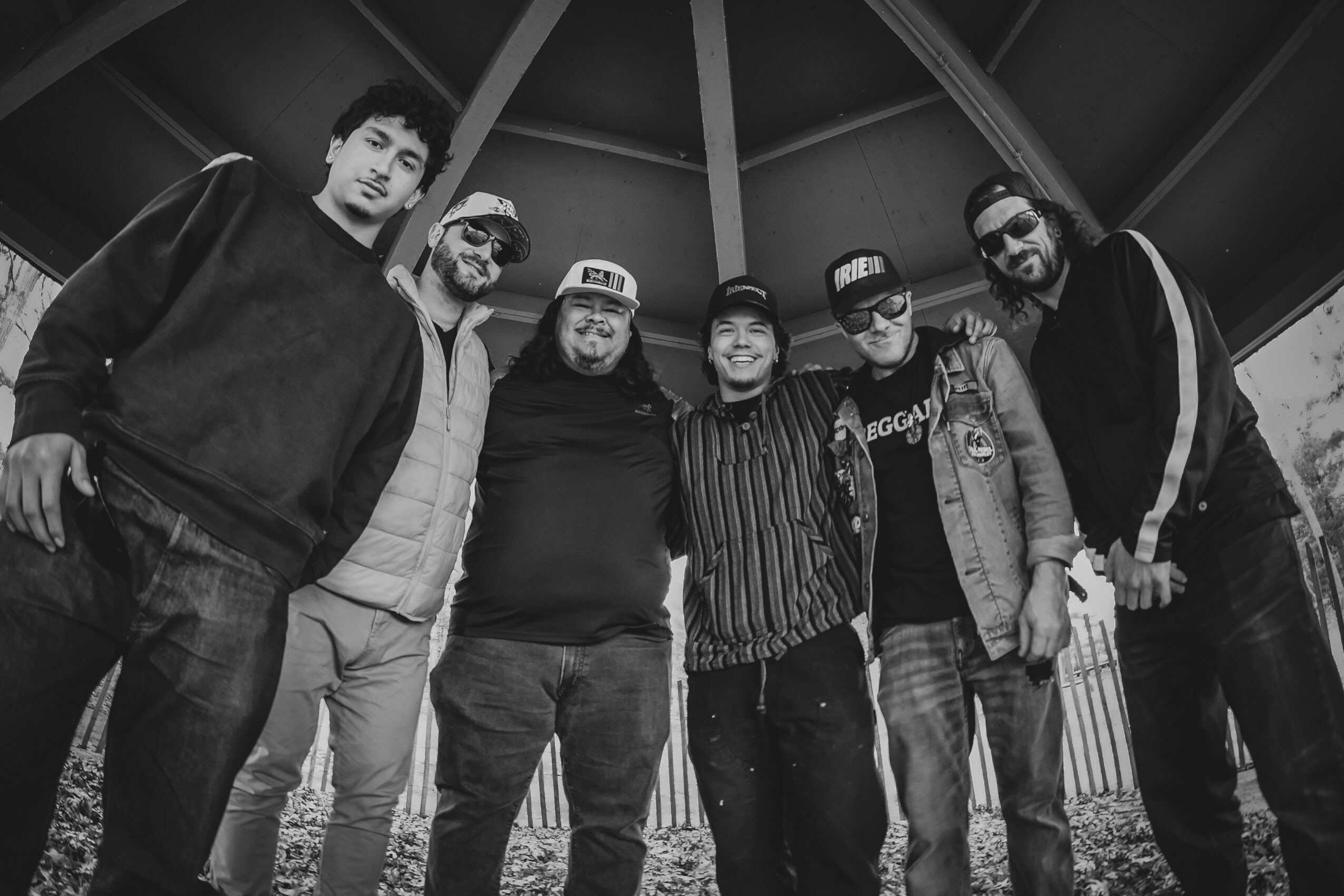 Group of seven people standing outdoors under a structure, smiling, wearing casual clothing and hats.