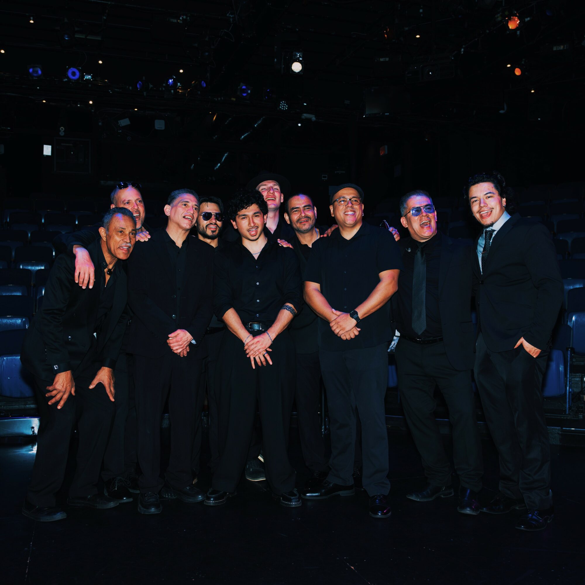 Group of nine people standing together in a dark setting, some wearing formal attire, smiling at the camera.
