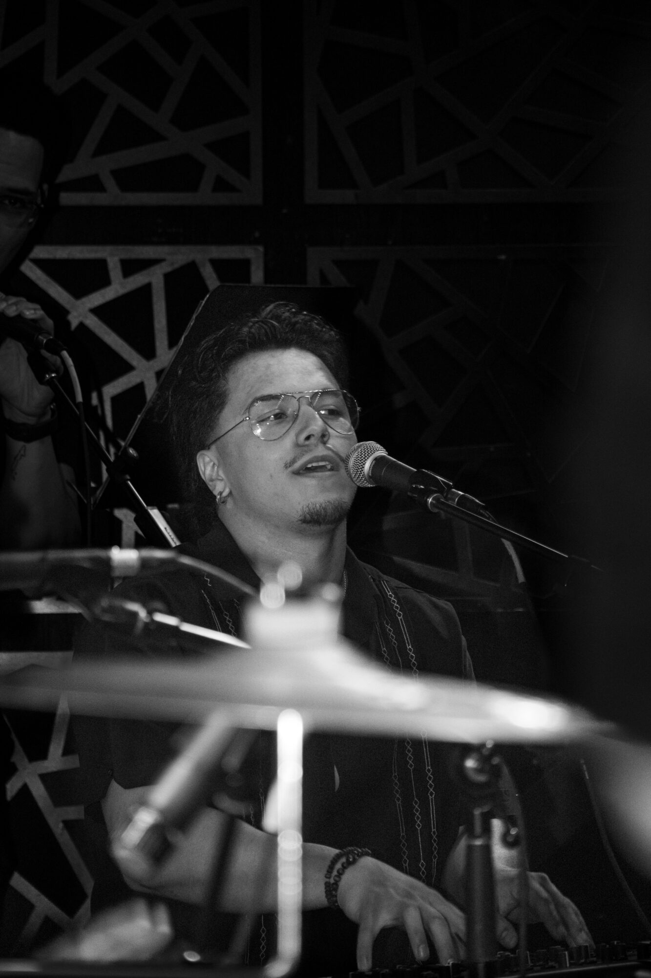 Person with glasses singing into a microphone during a performance, with musical equipment around.