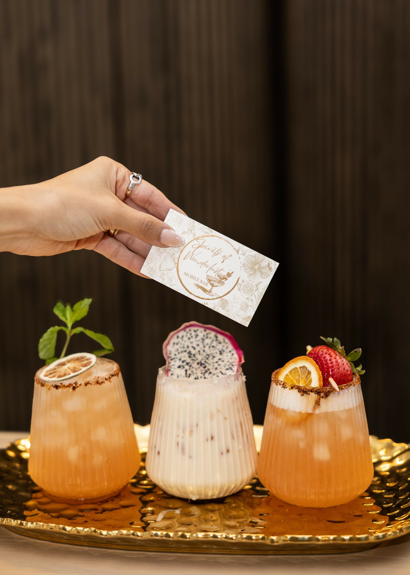 Hand holding a tea bag above three colorful drinks with fruit garnishes on a gold tray, wooden background.