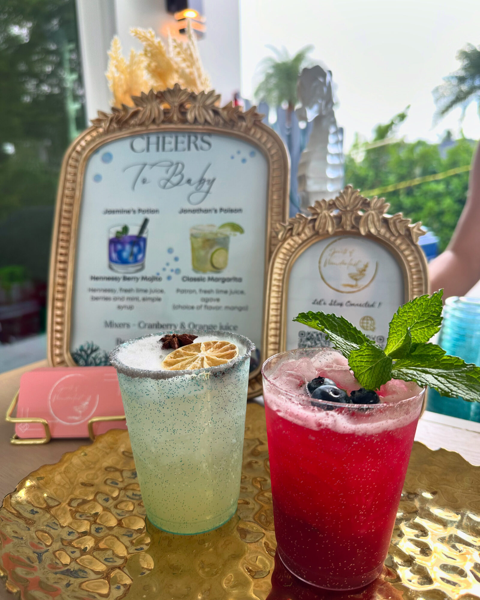Two colorful drinks on a gold table with framed menus and greenery in the background.
