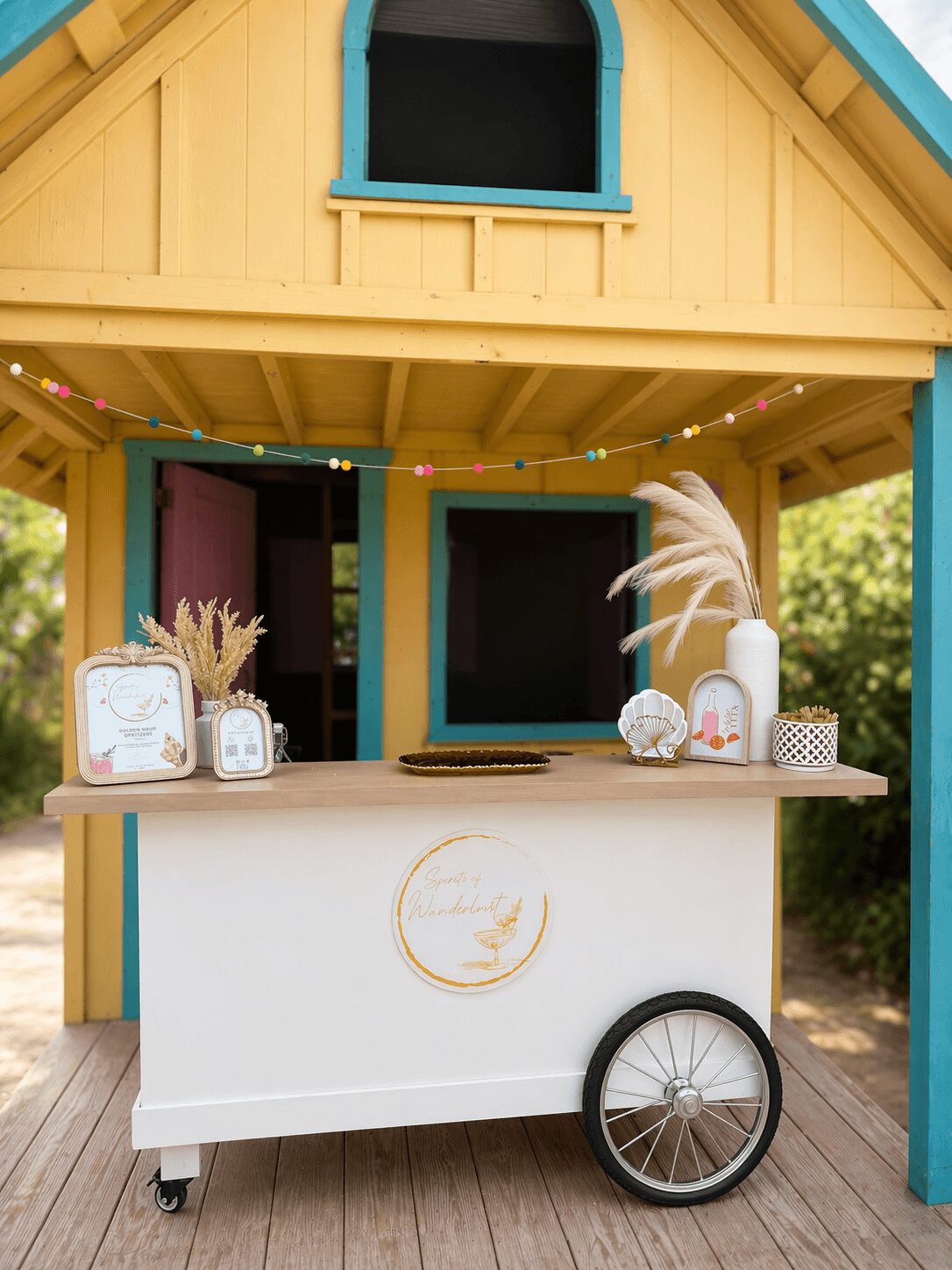 Colorful playhouse with a white cart in front, decorated with plants, crystals, and a feather, outdoors with trees in background.