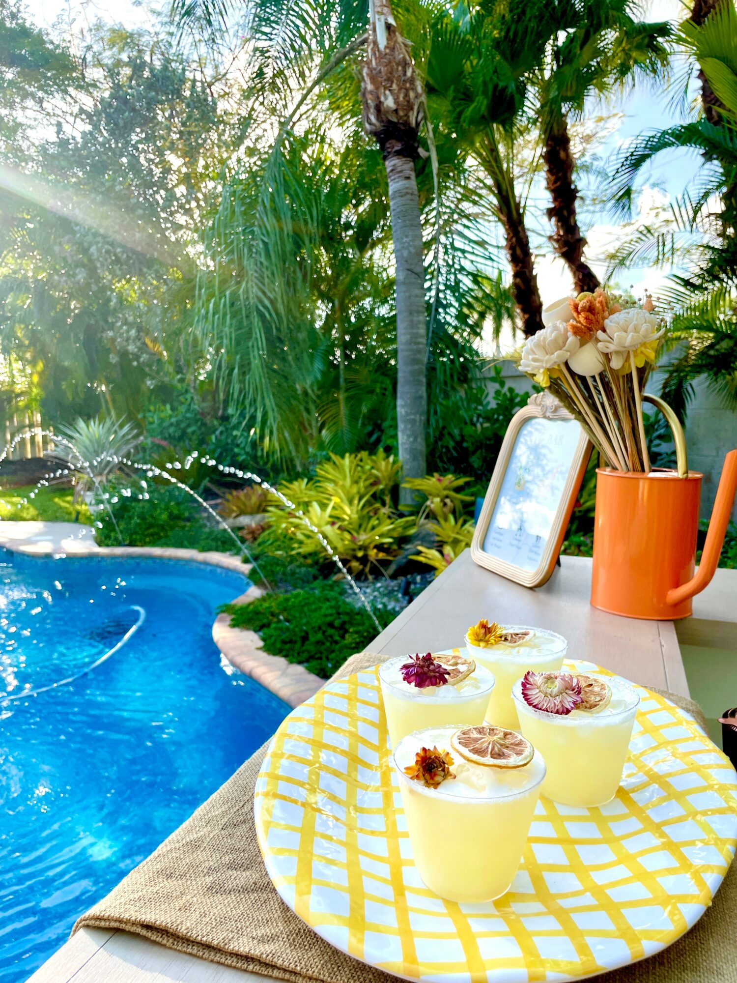 Poolside table with drinks, flowers, and a tablet, surrounded by lush tropical trees and plants.