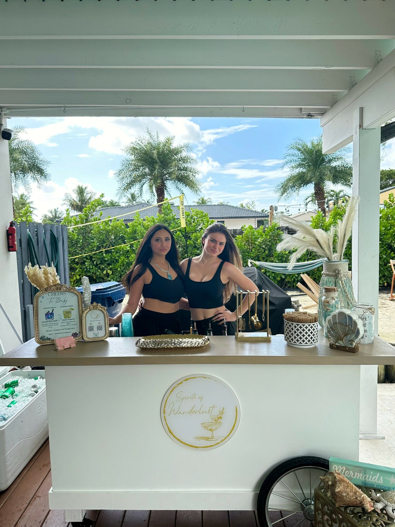 Two women stand behind a white counter with decorative items, outdoors with palm trees and blue sky in background.