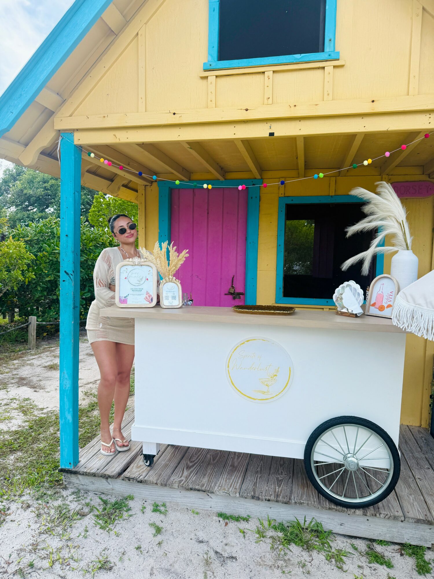 Person standing at a colorful outdoor stand with pink, yellow, and blue elements, wearing sunglasses and a beige outfit, with decorative feathers and items on the stand.