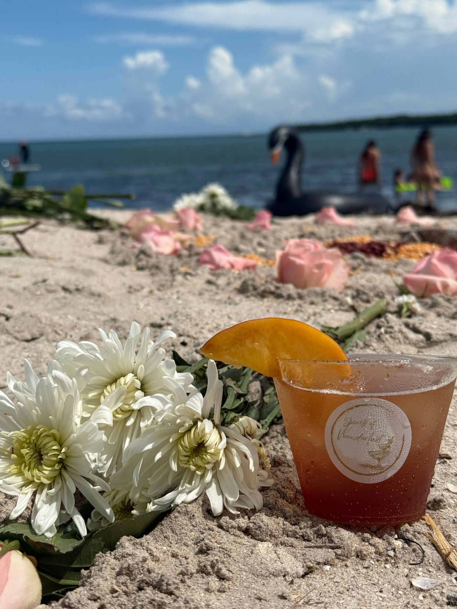 Beach scene with a drink, flowers, and people in the background, ocean, and sky, viewed from a low angle.