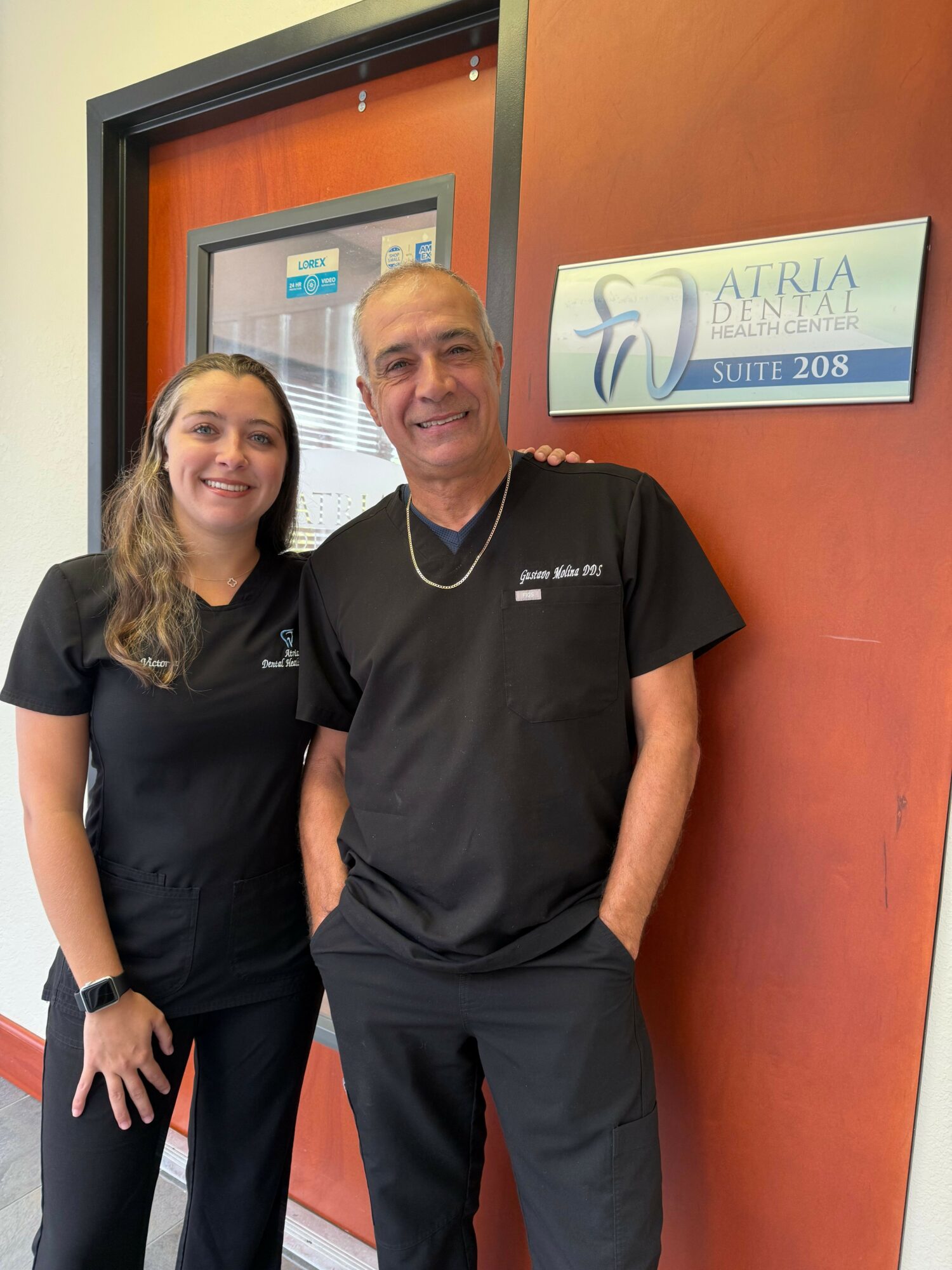 Two people standing in front of a door with a sign, smiling, one with short blonde hair, the other with long brown hair, both in black clothing.
