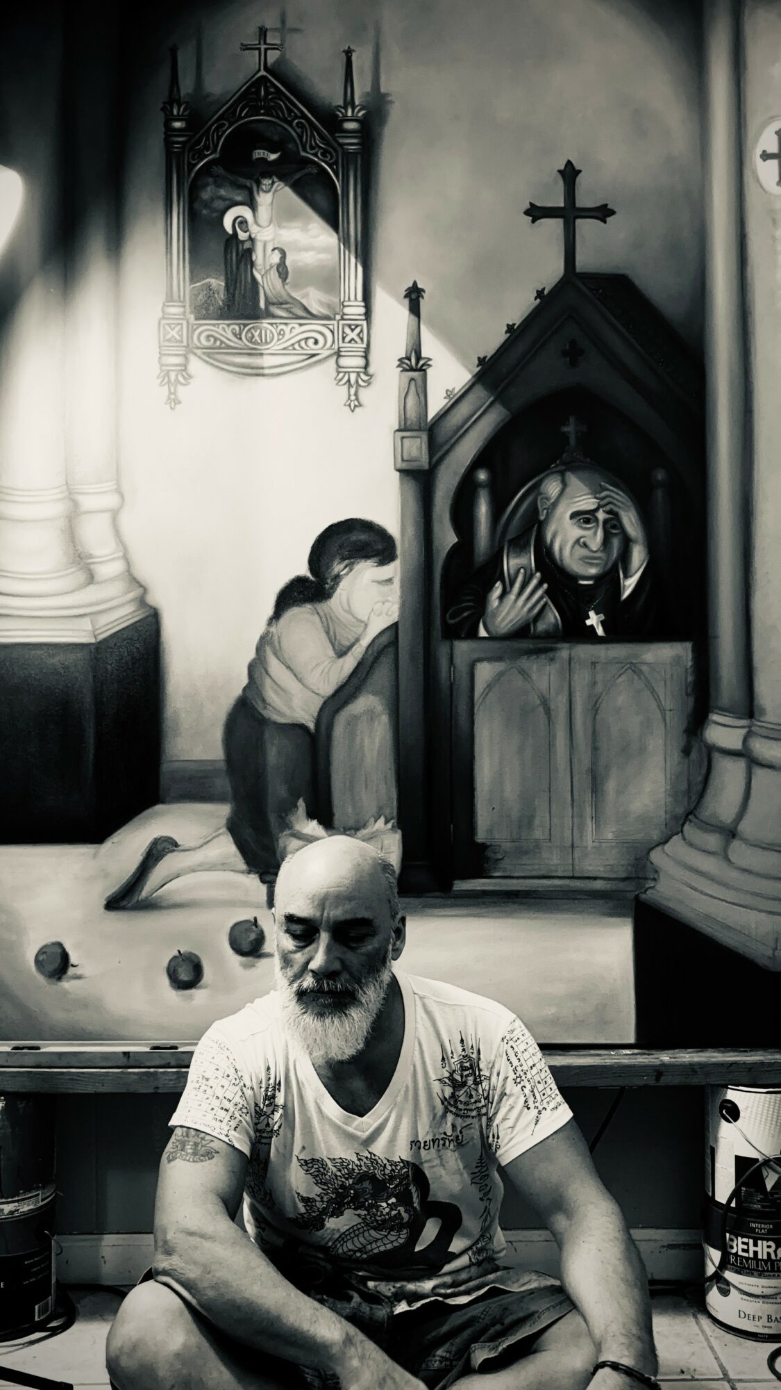 A man with a shaved head and tattoos sits in front of a religious scene inside a church, with a woman praying nearby.