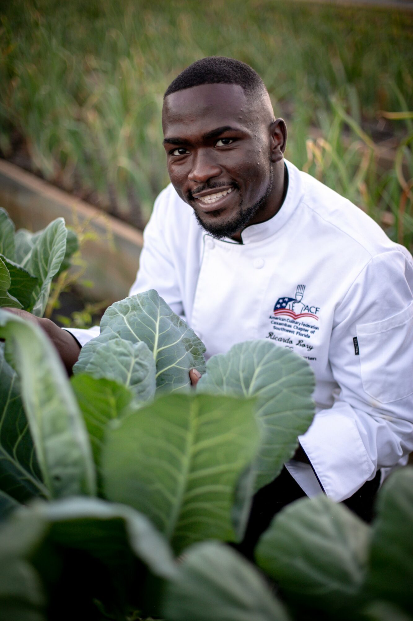 Man smiling in a garden with large leafy plants, wearing a white shirt with a logo.