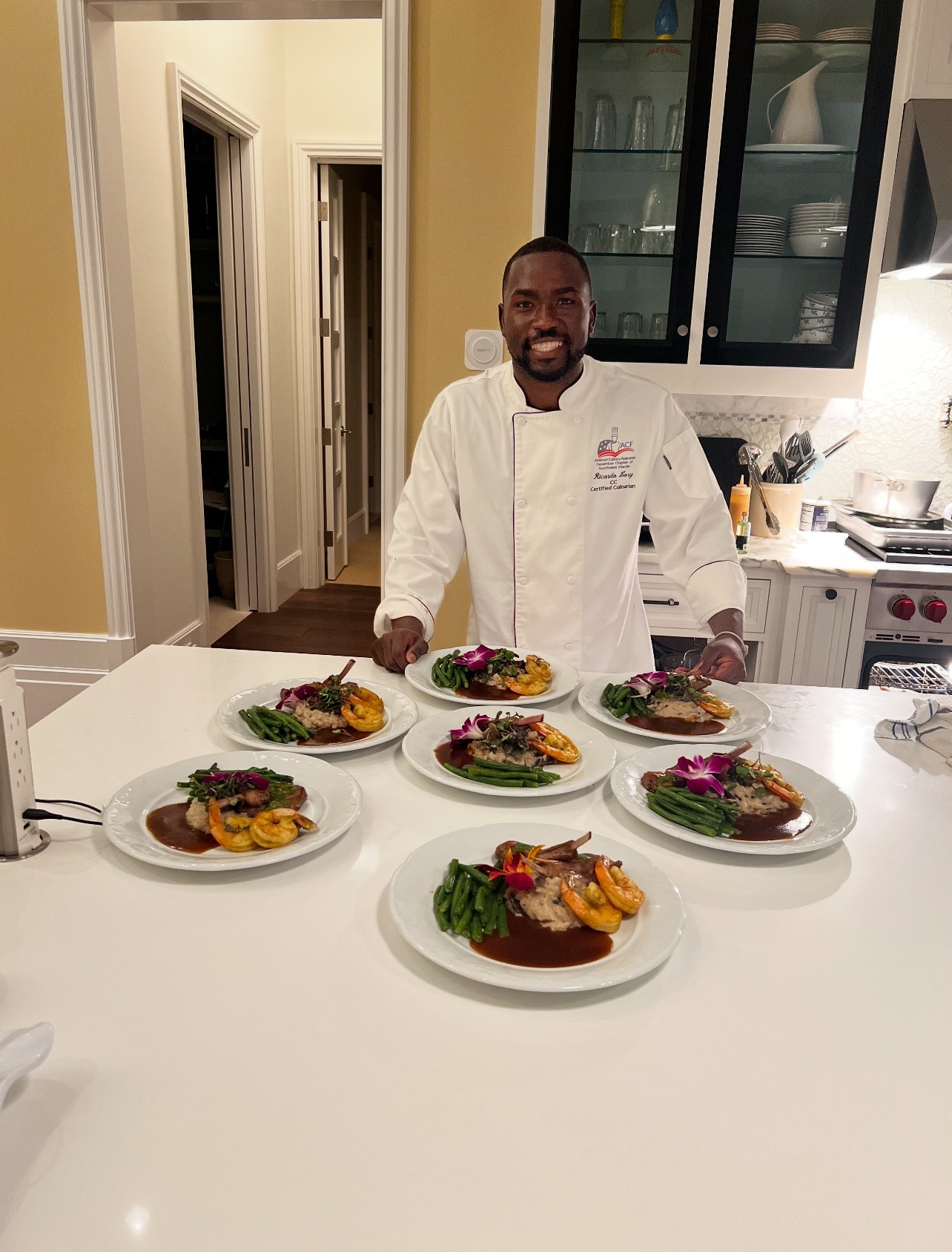 Chef standing behind a white kitchen island with six plated dishes, smiling, in a modern kitchen with glass cabinets.