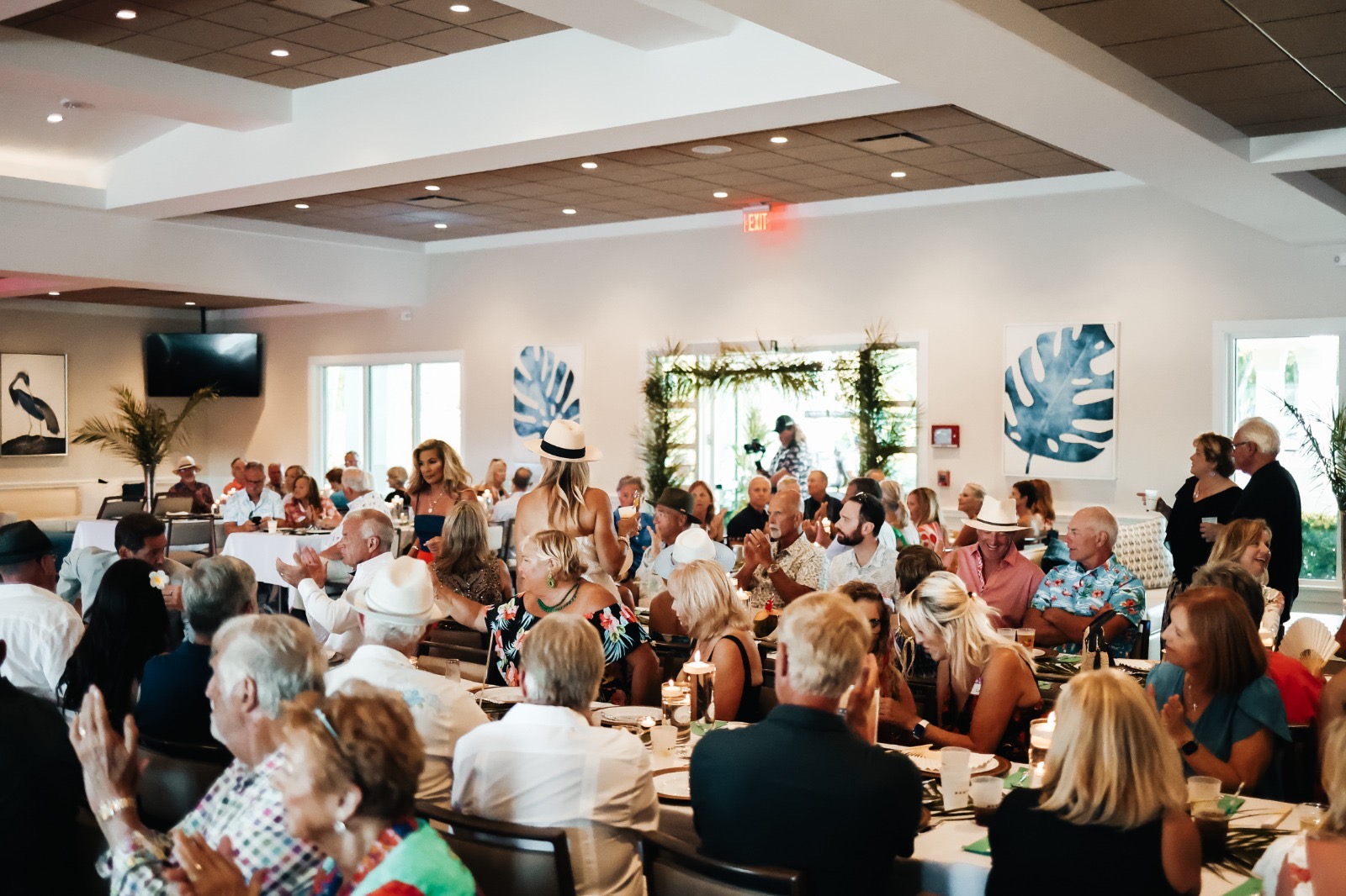 Large group of people seated at tables in a bright, modern room with tropical-themed wall art.