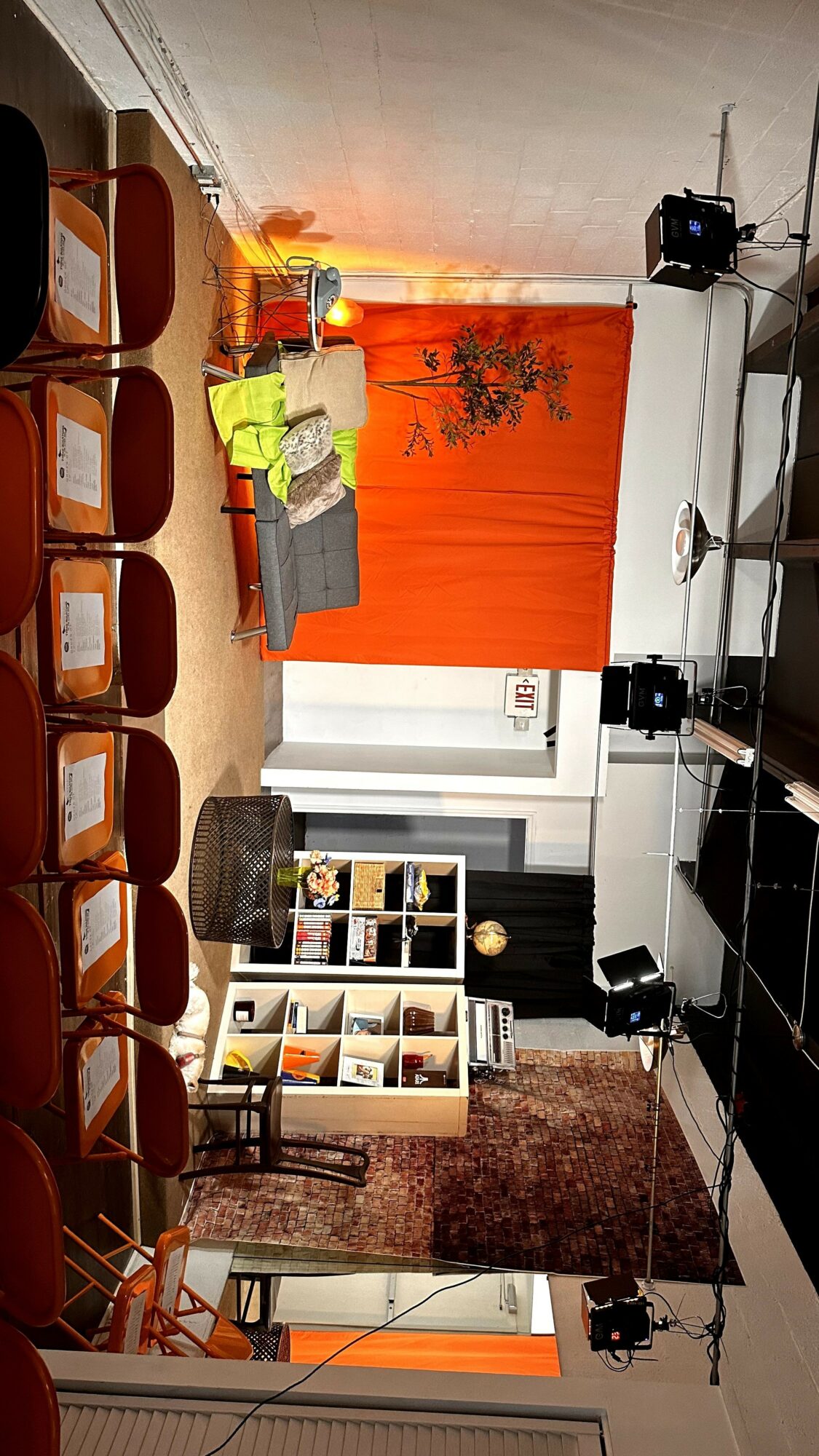 The stage has a gray sofa, colorful pillows, a bookshelf, and an orange backdrop with a tree silhouette, set for a performance.