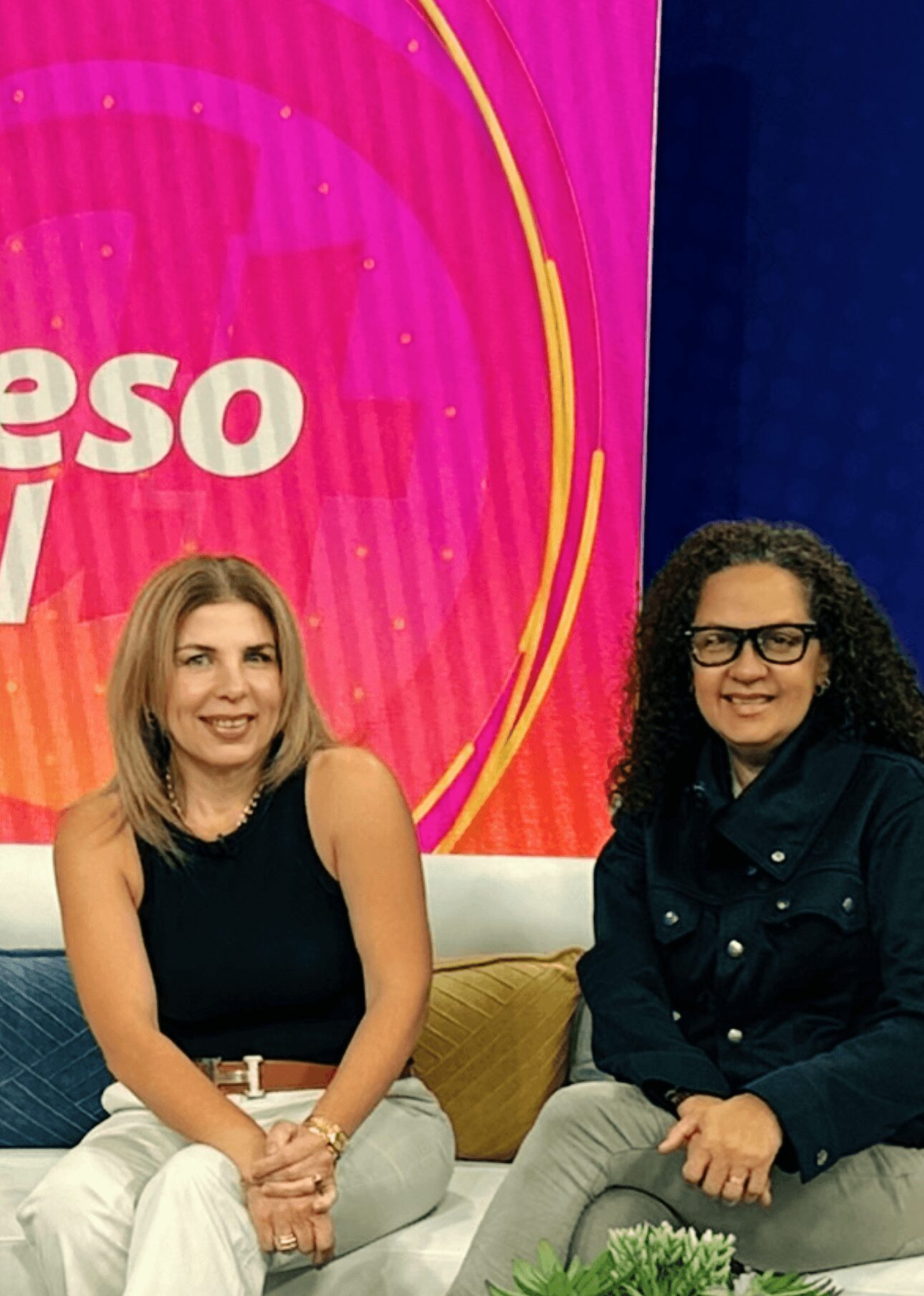 Two women sitting on a couch in front of a pink and blue background with text, smiling at the camera.