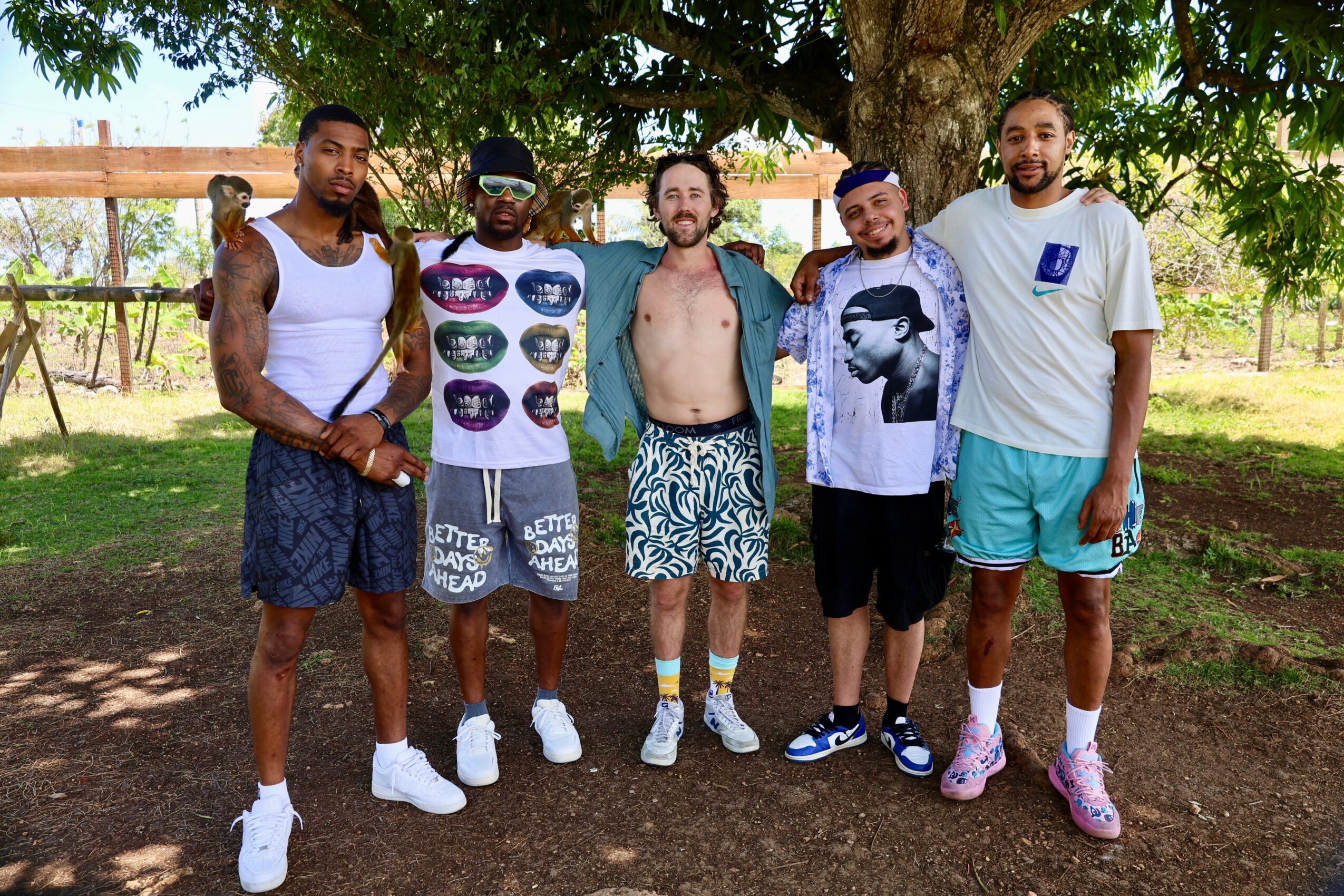 Five men stand outdoors under a tree, some wearing swimwear and casual clothing, smiling at the camera.